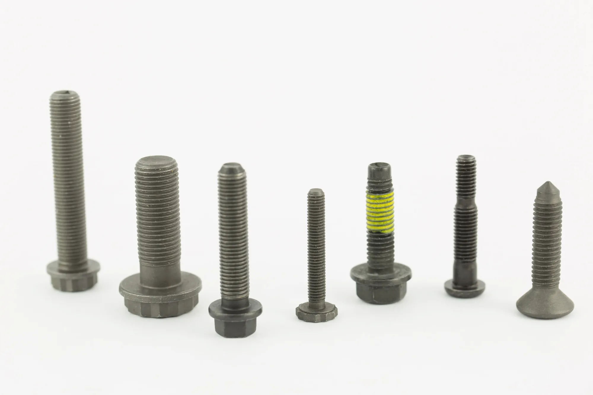 Seven automotive bolts standing upright on a white background: flanged hex, shoulder and countersunk head bolts in carbon steel with a plain black finish, one with a yellow thread-locking patch.
